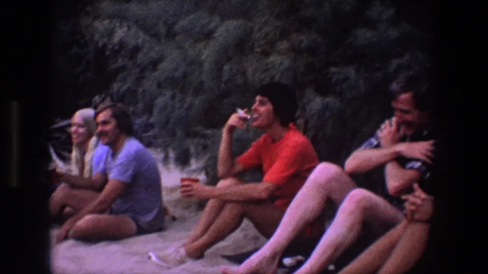 Young adults relax on a sandy Utah setting in 1969, including a man in a red shirt drinking while others sit nearby.
