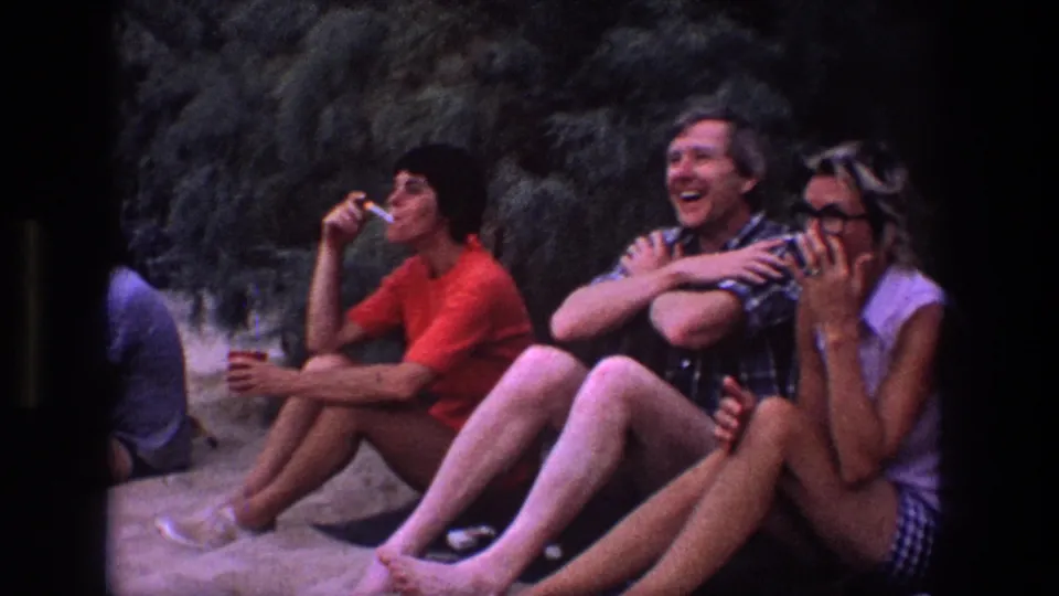 The frame shows friends in 1969 Utah home-movie footage laughing on sand, with one person smoking and others holding cups.