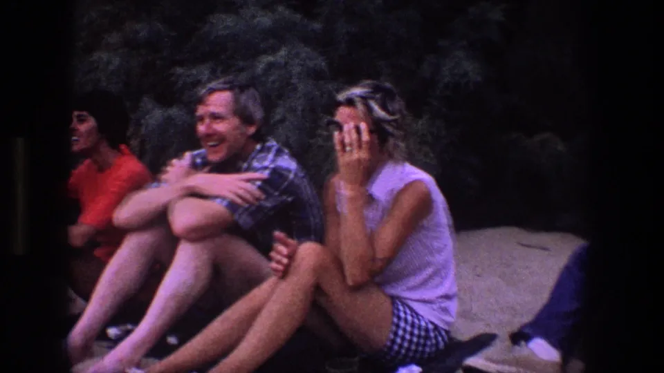 A tight 8mm frame shows three young adults seated on sand in Utah, smiling and talking beside dark wooded brush.