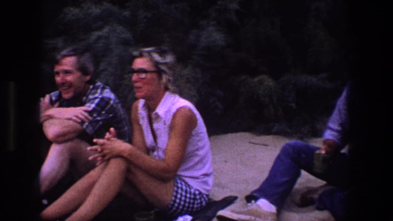 Young adults sit on sand near dark foliage, including a woman in glasses and a man in a patterned shirt.