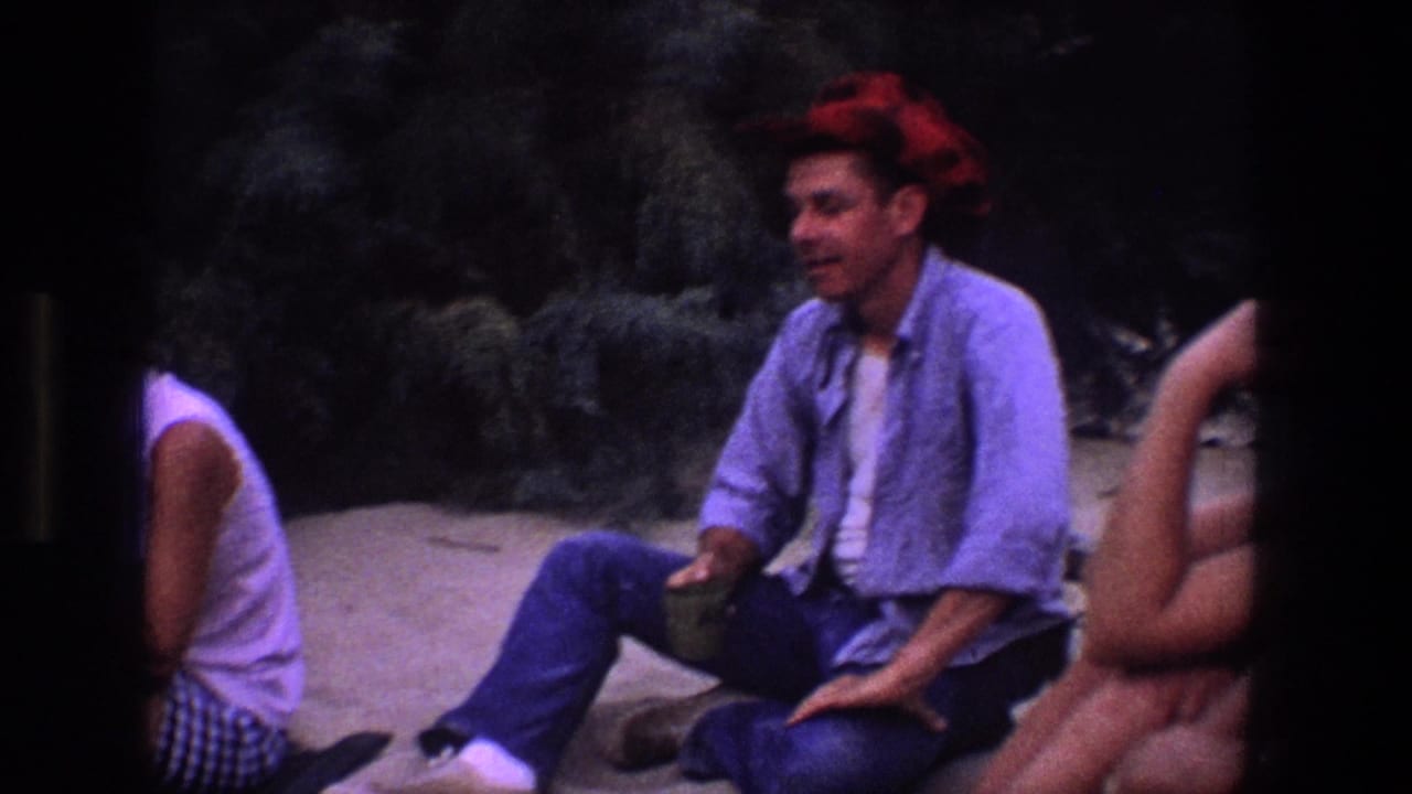 A man wearing a red bandana sits on sand beside partly visible companions and dark brush.