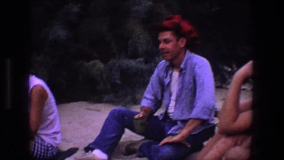 A man in a red bandana sits on sandy ground in the Utah home-movie footage, with other adults partly visible nearby.