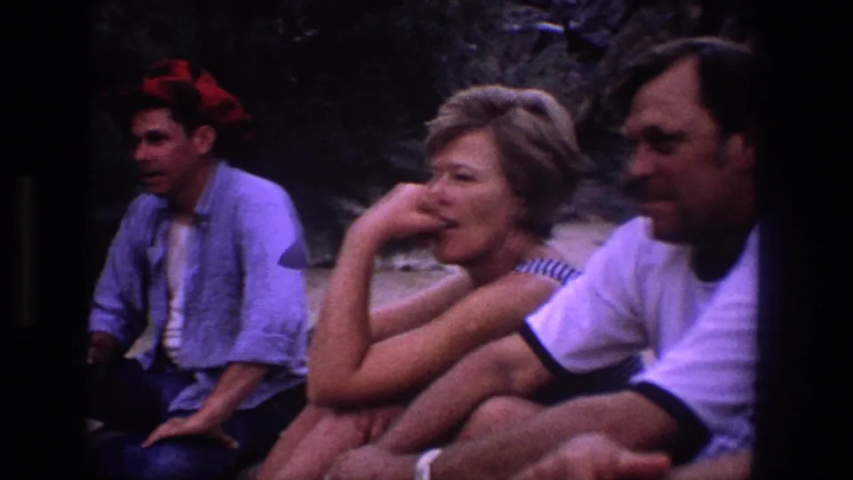 Adults sit close together on sand in the 1969 Utah footage, with faces visible and the handheld camera framing them tightly.