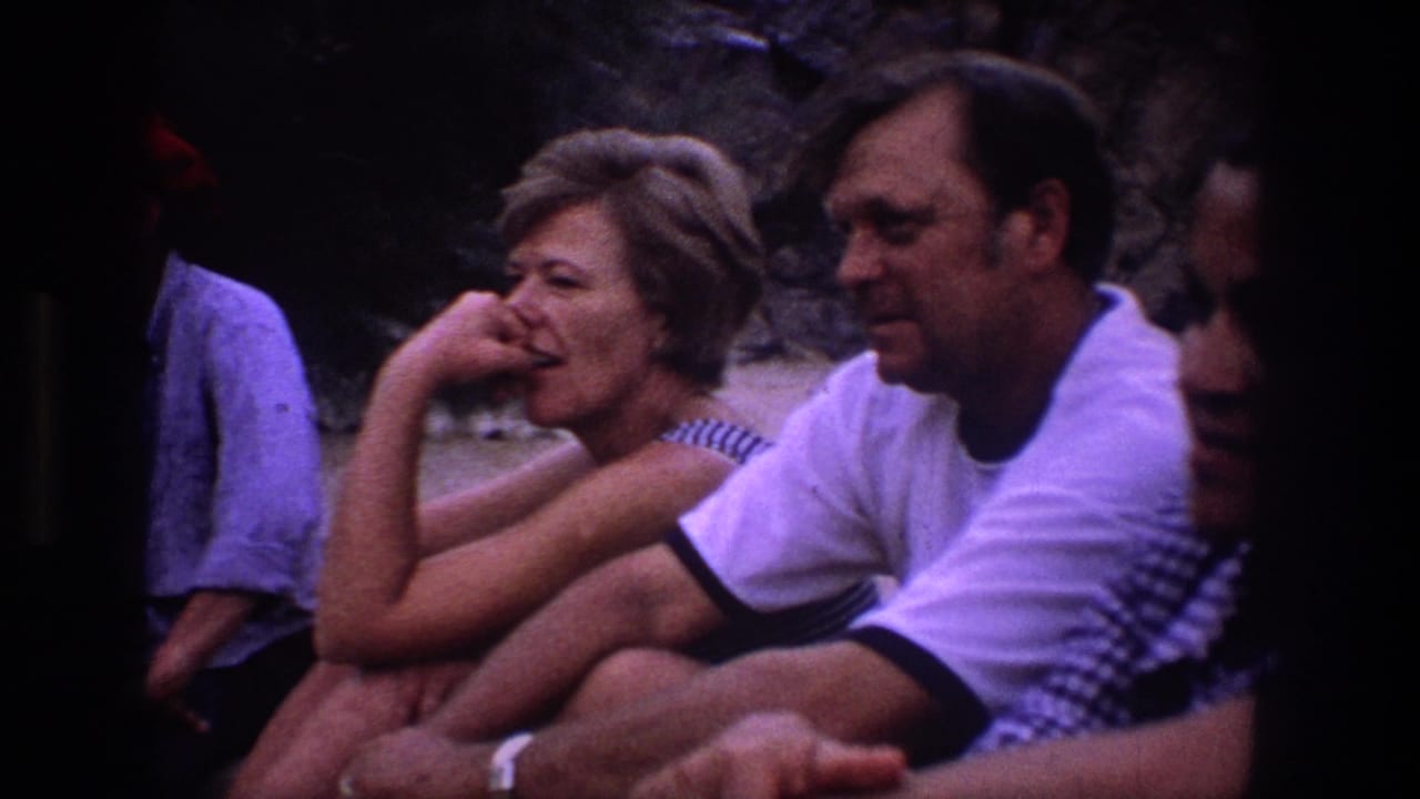 Utah 1969 home-movie frame showing adults seated on sand, with a woman in stripes and a man in a white T-shirt near a rocky background.