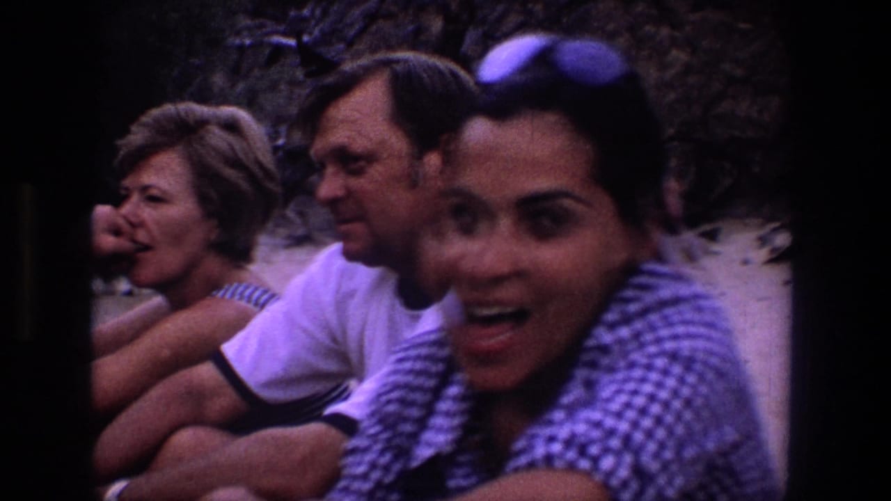 A smiling woman with sunglasses on her head leans near seated adults on the sand.