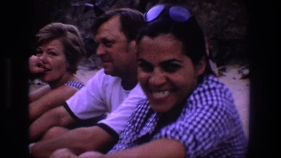 Utah 1969 home-movie frame showing a woman smiling in the foreground, with a man in a white T-shirt and a woman in stripes seated behind her.