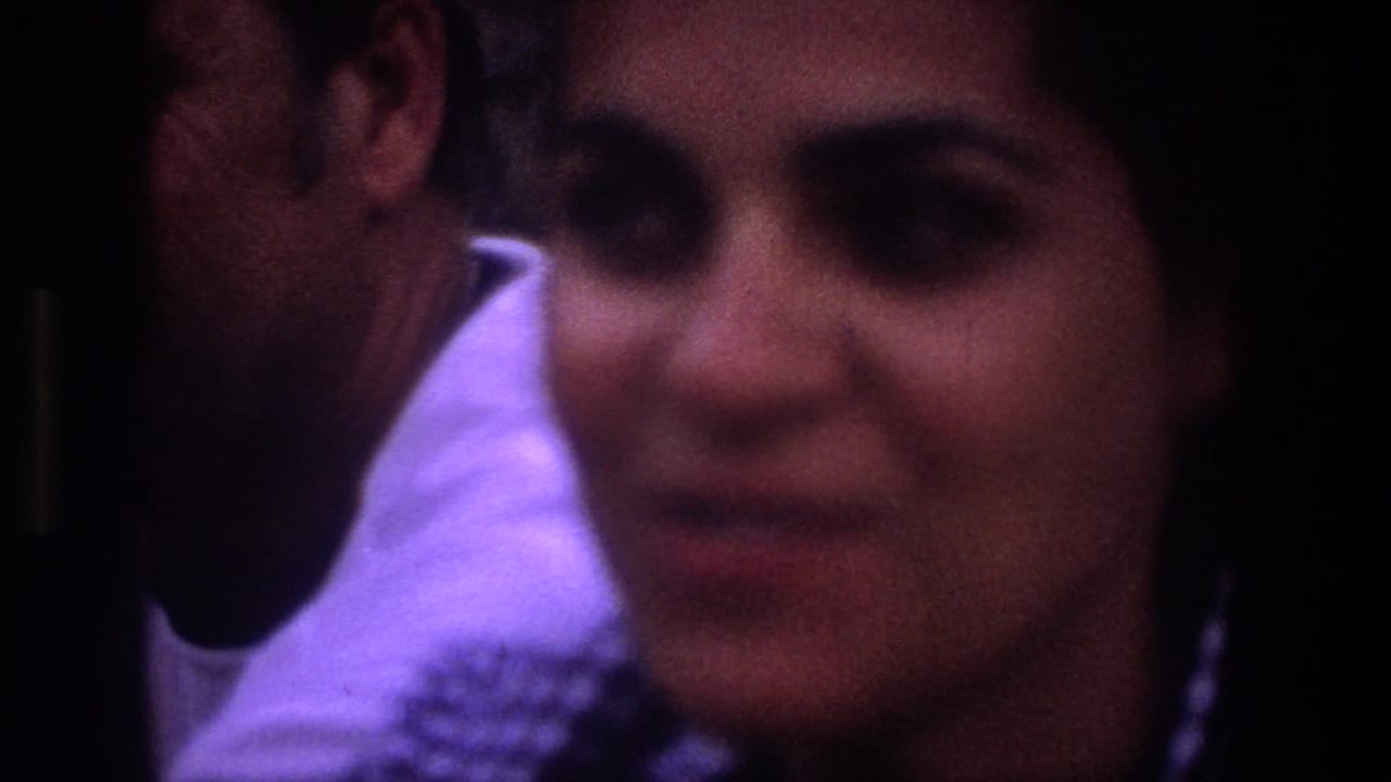 In 1969 Utah 8mm footage, a dark-haired woman looks toward the camera while a man sits close beside her in low purple light.
