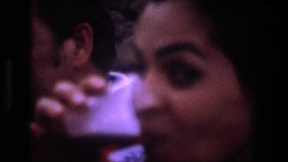 The woman lifts a small drinking glass toward the camera in the Utah 1969 home-movie scene, with a man partially visible beside her.