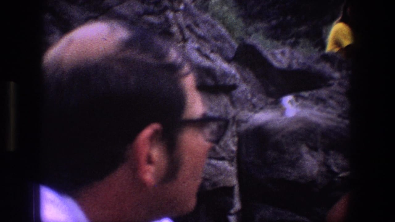 In Utah 1969 home-movie footage, a man in dark glasses looks toward a rocky cliff face where a small pale bird is visible.