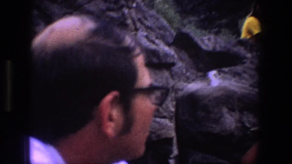 In Utah 1969 home-movie footage, a man in dark glasses looks toward a rocky cliff face where a small pale bird is visible.