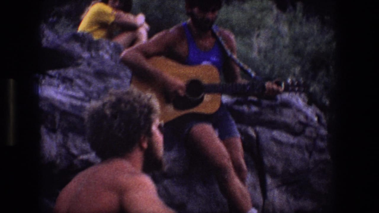 Man in a blue tank top plays acoustic guitar on rocks while friends sit nearby outdoors.