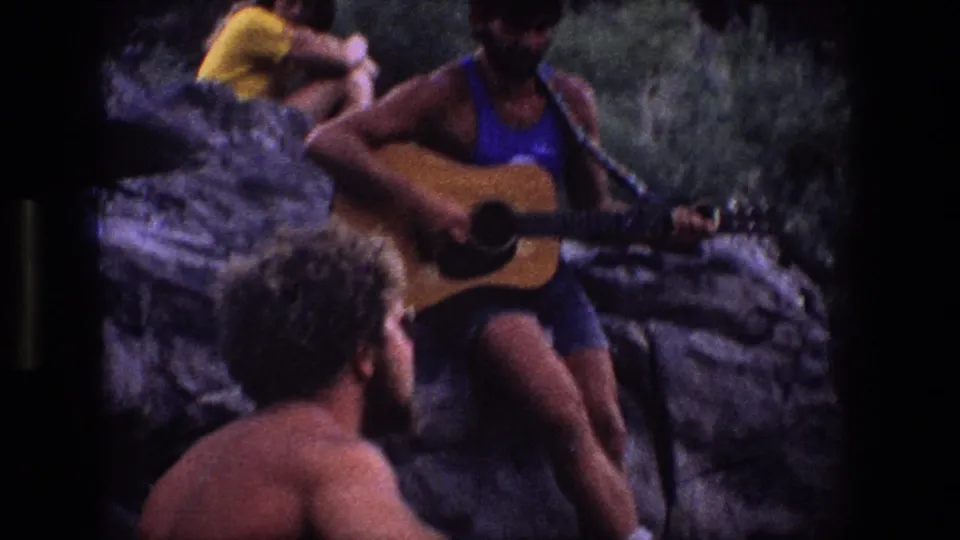 In 1969 Utah home-movie footage, a man plays acoustic guitar on a rocky outcrop as a woman and another listener sit nearby.