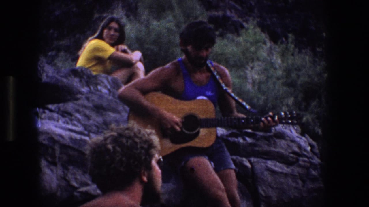 A man with an acoustic guitar sits on rocks in a Utah outdoor setting while a woman in yellow and a foreground listener watch.