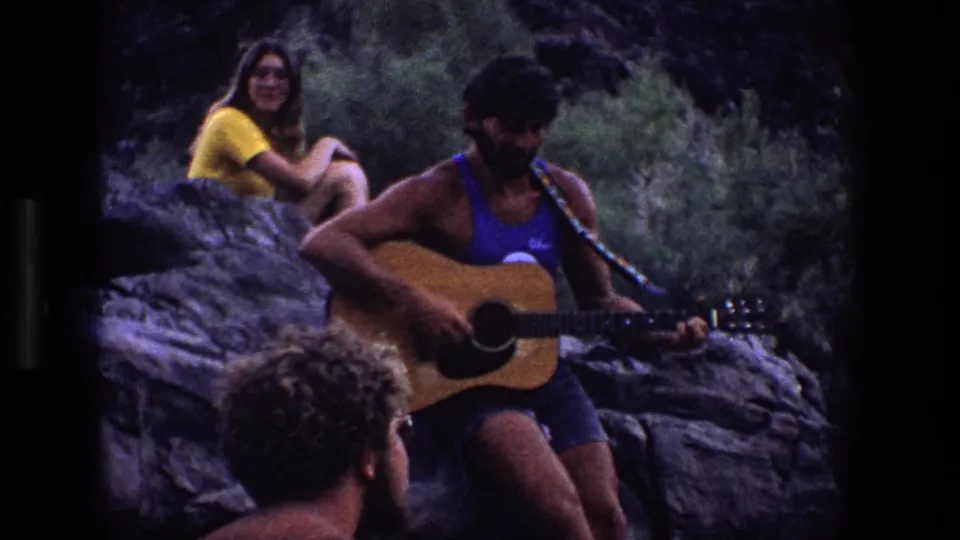 The woman in yellow smiles as the guitar player sits below her on rocks, with a curly-haired listener visible in the foreground.