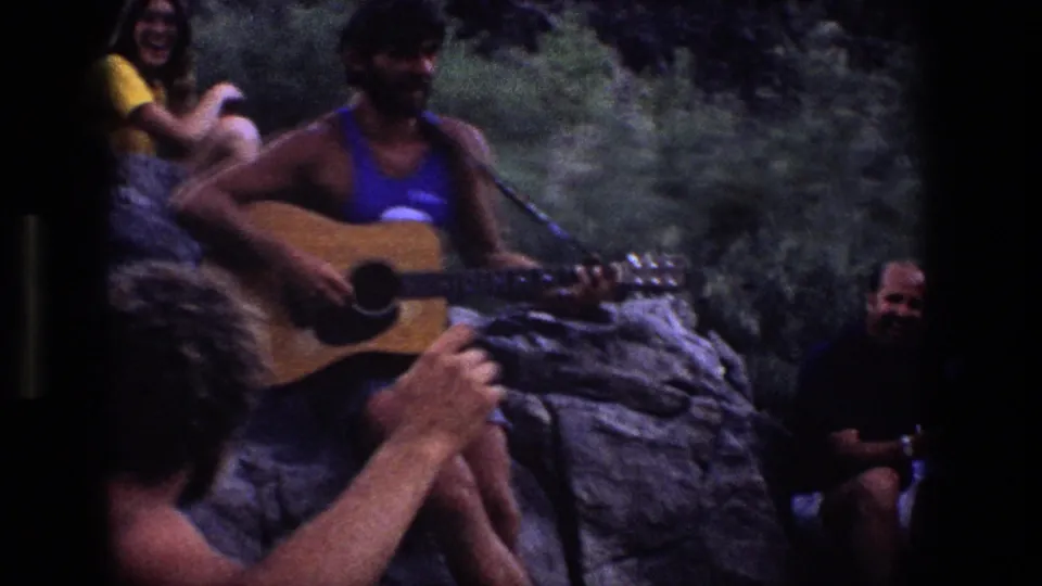 A closer 8mm frame shows the guitar player seated on rocks, with a smiling woman behind him and another seated man partly visible at right.