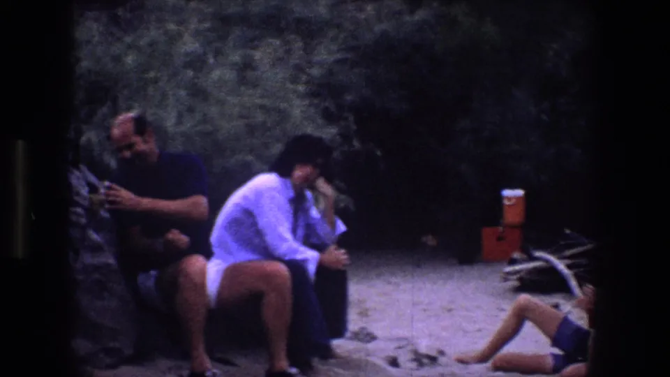 1969 Utah 8mm frame with adults sitting against a rocky backdrop, a portable cooler on sand, and a partly visible seated figure at the right edge.