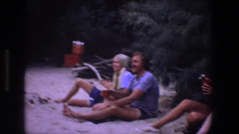1969 Utah 8mm frame showing friends seated barefoot on sand near a portable cooler, looking toward activity outside the frame.