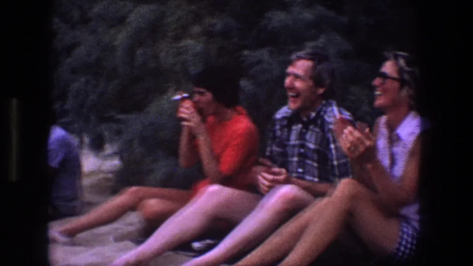 1969 Utah 8mm frame of adults sitting close together on sand, smiling and laughing, with one person holding a small camera.
