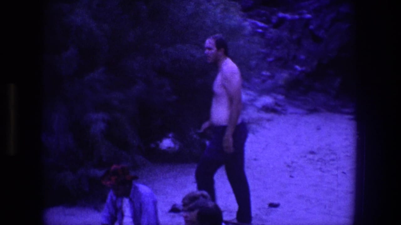 Shirtless man in dark pants stands on a rocky path with bags nearby.