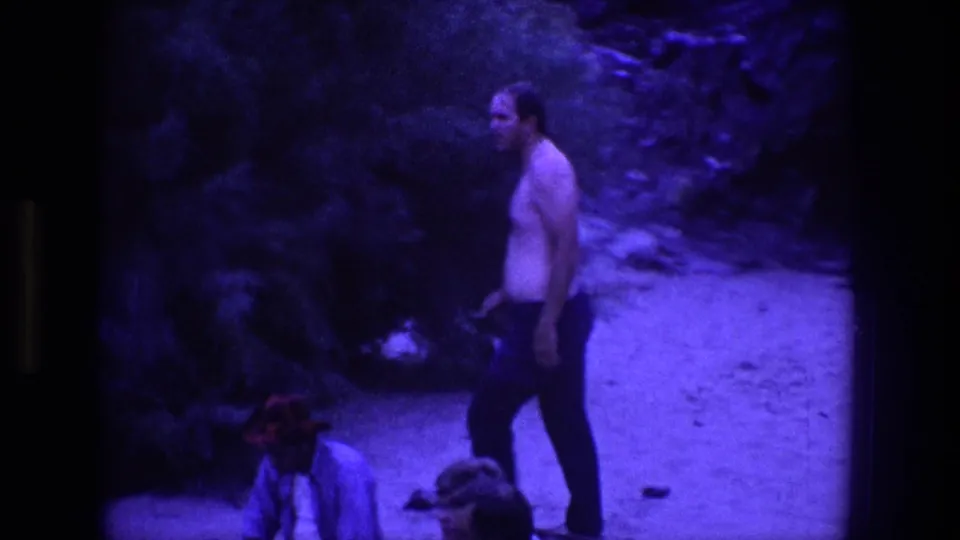 A shirtless man in dark pants stands on a rough Utah path, with rocky ground, shrubs, and personal items visible near him.