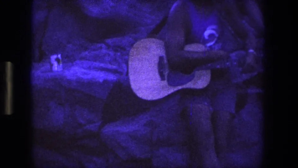 The guitar body and the seated player's upper body are visible against rough stone under intense purple-blue lighting.
