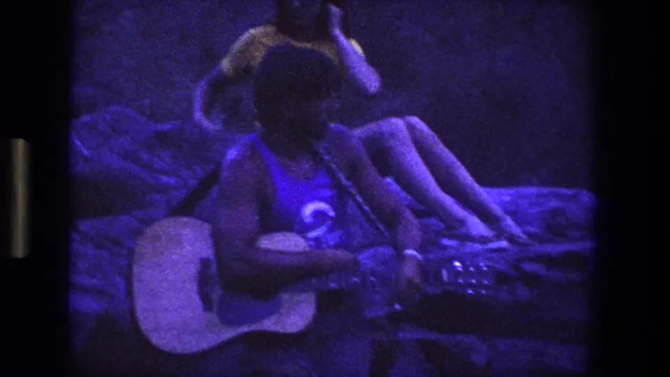 A curly-haired young man wearing glasses sits with an acoustic guitar in a rocky Utah setting, seen through grainy 8mm footage.