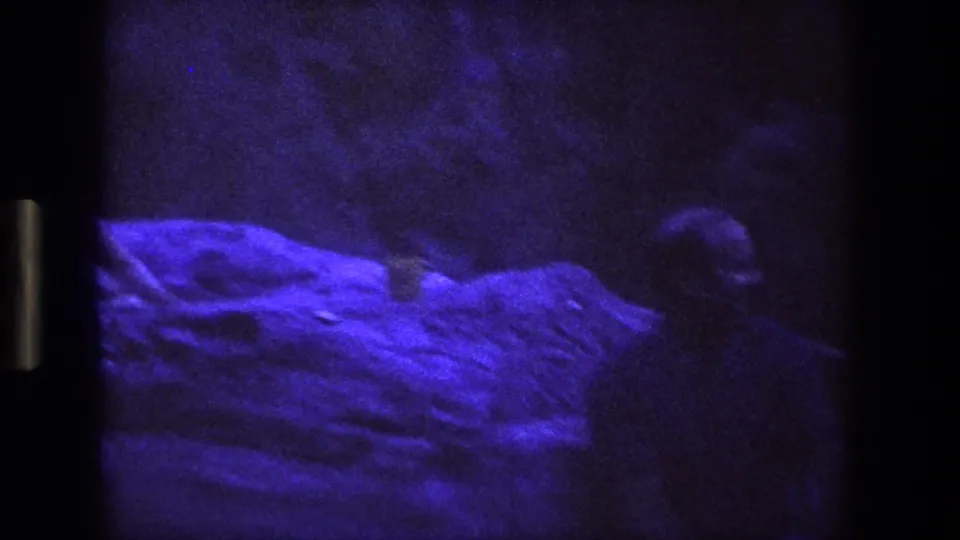 A dark, purple-lit rocky alcove shows the guitarist partly at the edge of frame, with the stone surface dominating the view.
