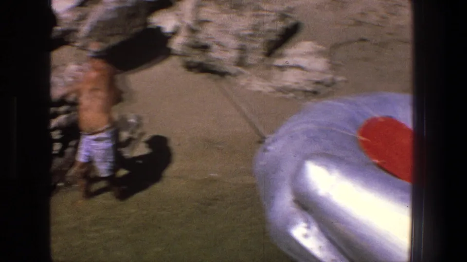The camera view shifts to a shirtless man in plaid swim trunks standing near rocks and sand along the Utah shoreline.