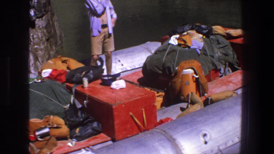 The frame shifts to the loaded raft and a partly visible standing man, with bedding, bags, and a red box near the canyon wall.