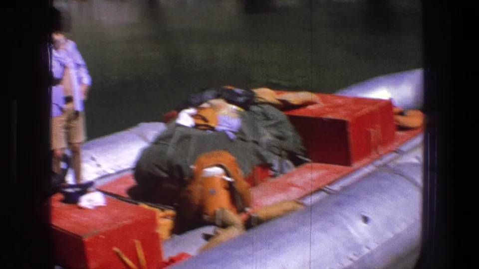 1969 Utah river scene with a woman watching a gray inflatable raft carrying red boxes, bedding, and camping equipment.