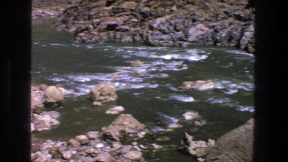 1969 Utah home-movie frame of whitewater moving through a rocky river channel beside the edge of a loaded raft.