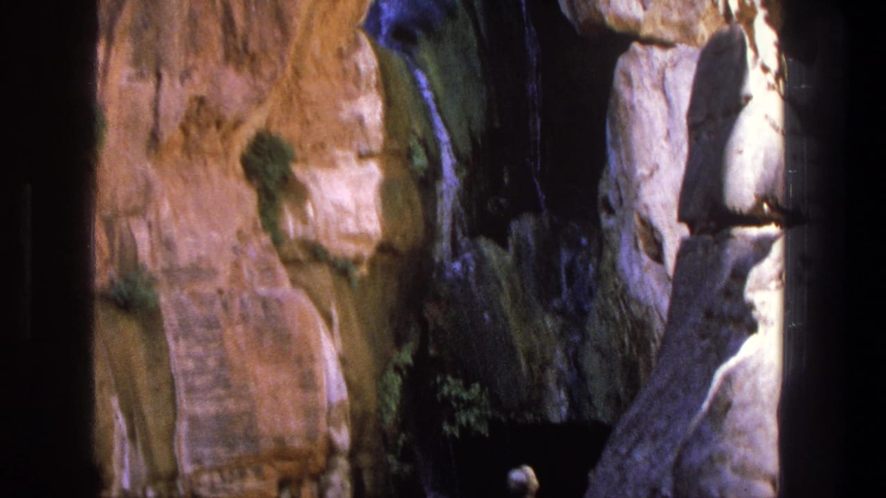 Tan canyon wall with a small waterfall and a few people near shallow water.