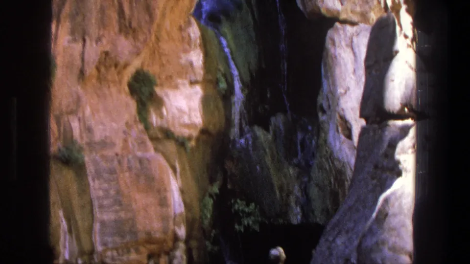 1969 Utah 8mm frame showing tan canyon rock, a dark recess, a thin waterfall, and visitors near a shallow natural pool.
