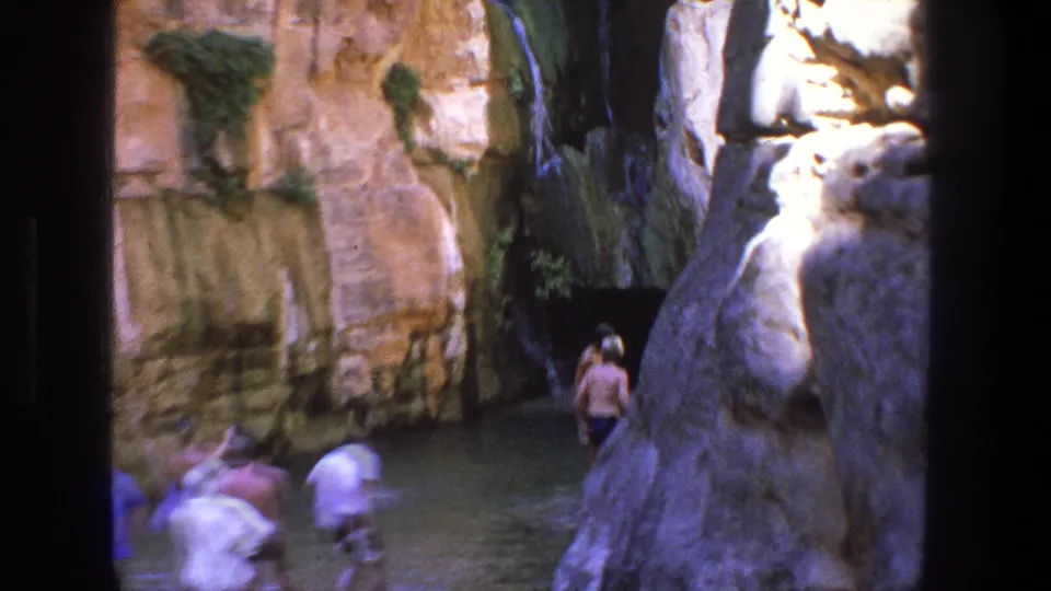 1969 Utah home-movie frame with several visitors wading or walking through a narrow canyon pool below high rock walls.