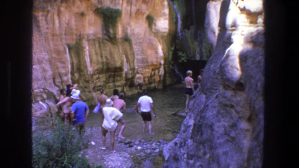 1969 Utah 8mm frame showing a small group gathered beside shallow water, with tan cliffs, rock ledges, and canyon shade behind them.
