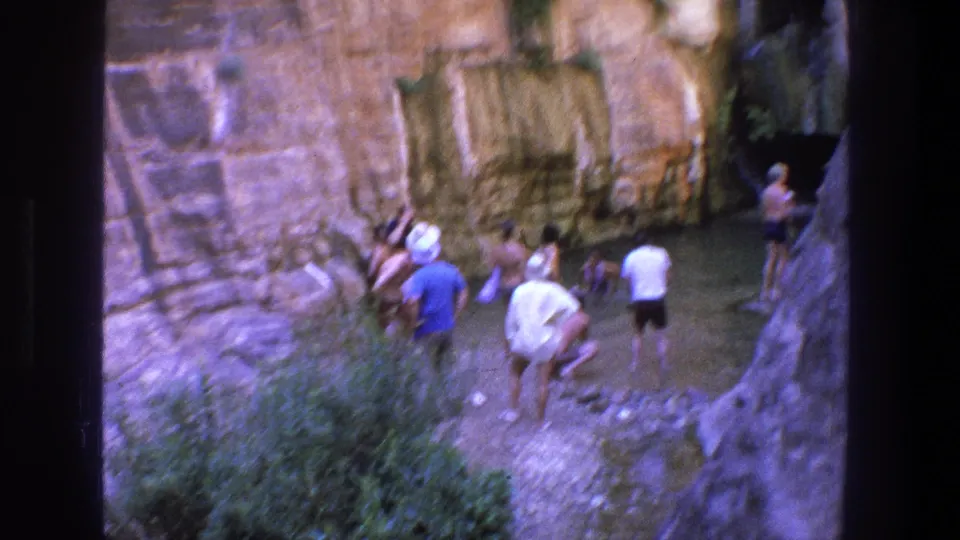 1969 Utah home-movie frame showing visitors around a shallow canyon pool, with blurred motion, tan stone walls, and foreground brush.