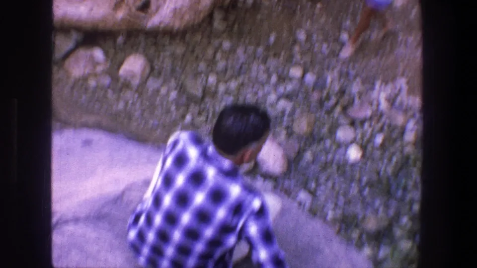 A man in a plaid shirt sits or crouches on pale rock beside a rough stone wall in the Utah home-movie scene.