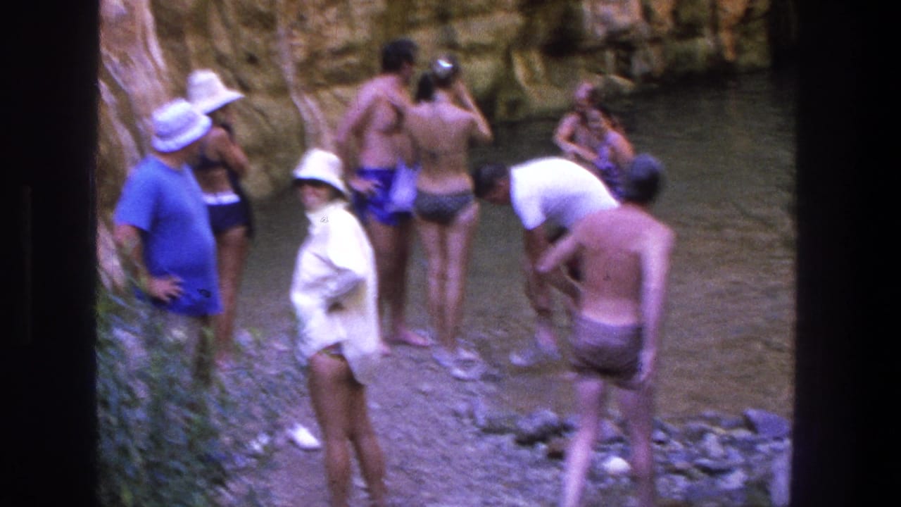 People in swimsuits and sun hats stand beside a shallow rocky creek pool.