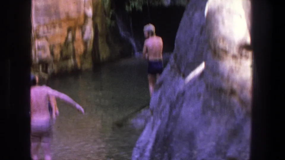 A home-movie view of swimmers entering or crossing a narrow Utah canyon pool, with bright rock and shaded water.