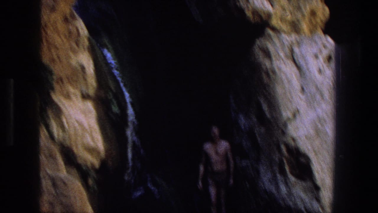 Man in swim trunks standing inside a dark rocky opening above water.
