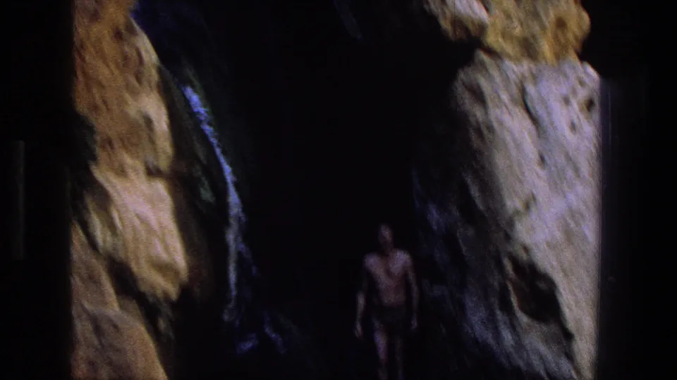 A man in swim trunks appears at a dark cave-like rock opening in the Utah canyon footage.