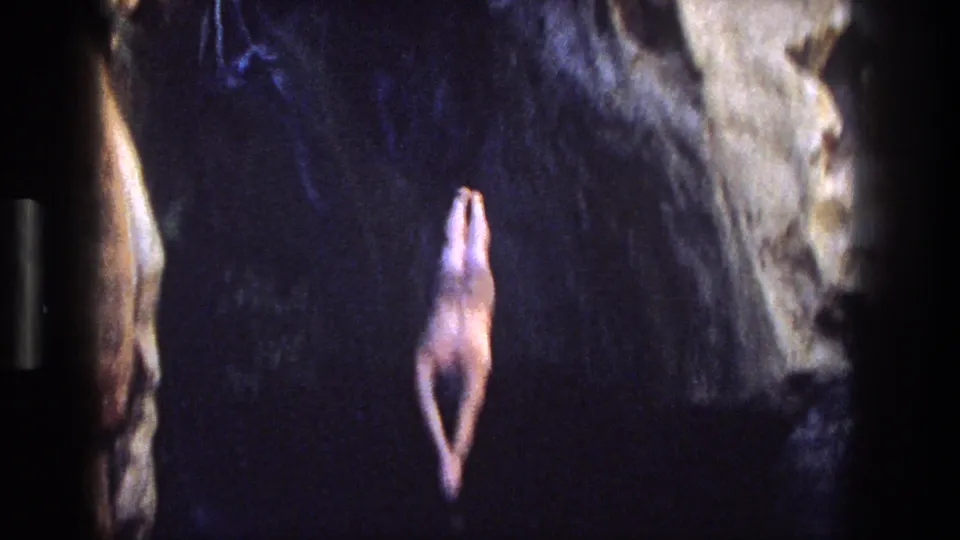 The man is captured mid-dive from a shadowed rock opening toward the natural pool below.