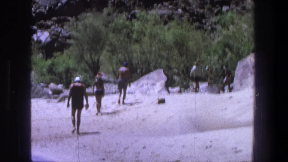 Three beachgoers walk away across tan sand toward green shrubs and rocky terrain in the Utah 1969 footage.
