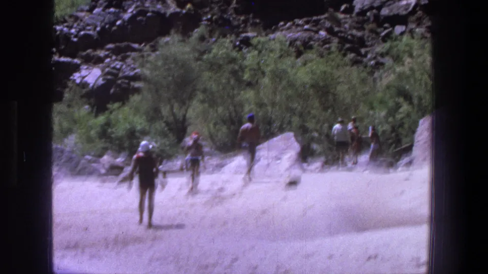 Beachgoers move across a sandy Utah recreation area with green shrubs, boulders and rocky canyon terrain behind them.