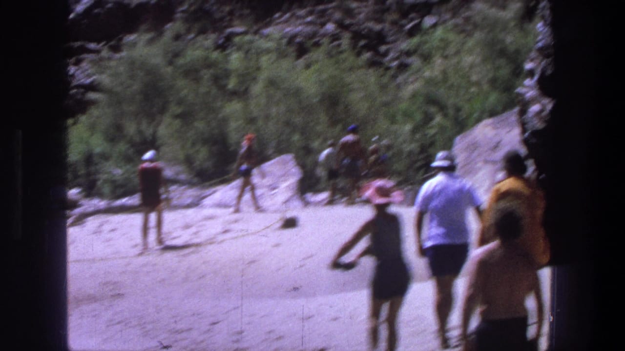 1969 Utah home-movie frame showing adults in vintage swimwear walking on a sunny sandy riverbank below rocky terrain.