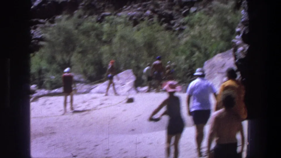 1969 Utah home-movie frame showing adults in vintage swimwear walking on a sunny sandy riverbank below rocky terrain.