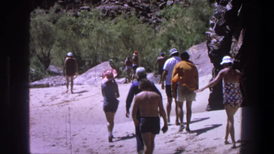 1969 Utah home-movie frame with several adults in vintage swimwear walking together along a sandy riverbank path.