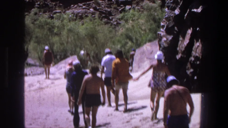 1969 Utah frame showing a casual group walking away on a sunlit riverbank, with rocks, shrubs, and shaded frame edges.