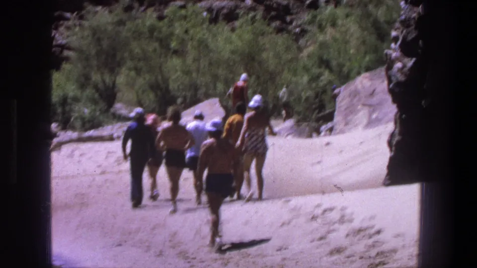 1969 Utah 8mm frame of adults in swimwear and hats walking away on a sandy path near rocks and brush.