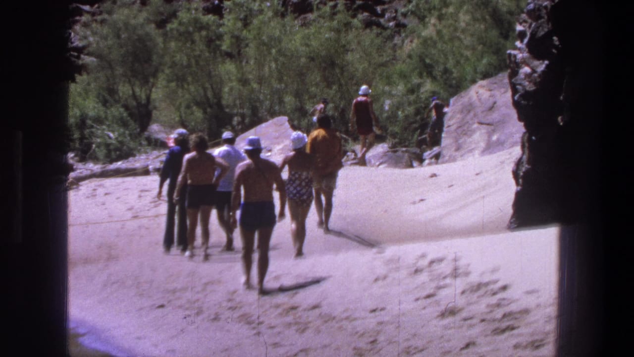 1969 Utah home-movie still showing a group in vintage swimwear moving along a sandy route by rocks and shrubs.